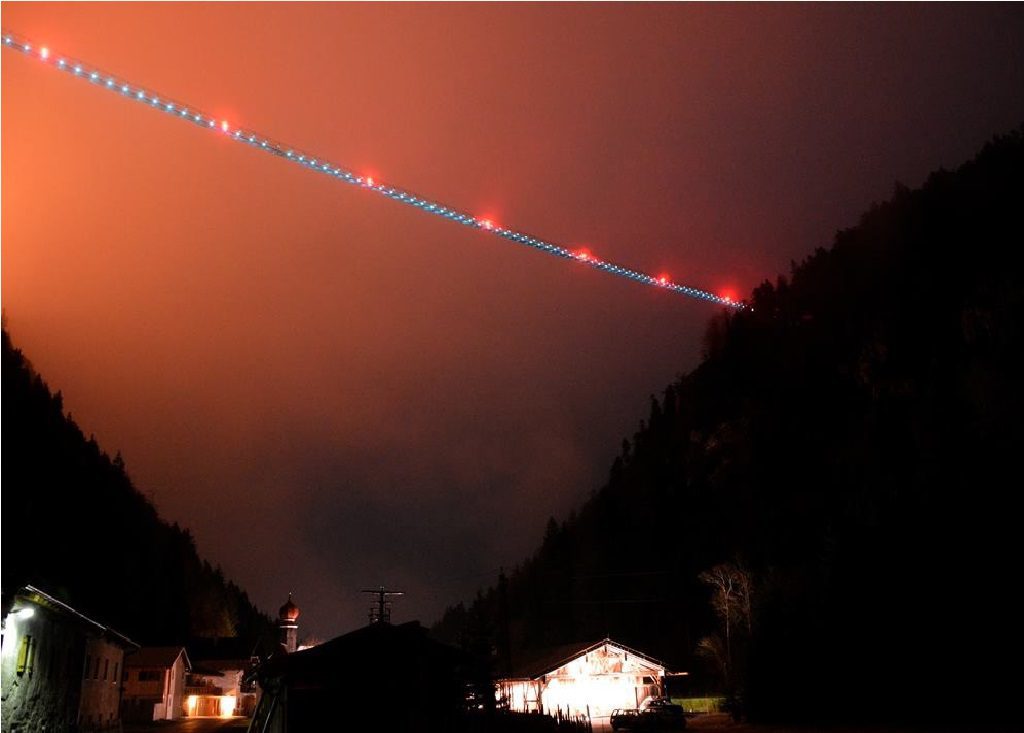 Długi, oświetlony most wiszący rozciągający się nad doliną, widoczny w nocy na tle pomarańczowo-czerwonego nieba. Poniżej, w ciemności, zarys domów z częściowo rozświetlonymi oknami i sylwetki gór porośniętych lasem.