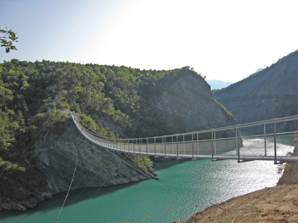 Długi, metalowy most wiszący rozciągający się nad turkusowym jeziorem, otoczony zalesionymi, skalistymi górami, pod jasnym niebem.