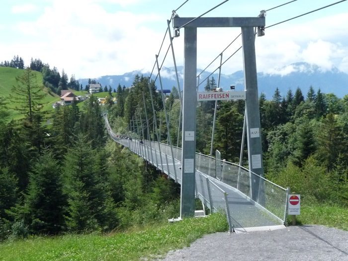 Długi, metalowy most wiszący Skywalk Raiffeisen rozciąga się nad zieloną doliną porośniętą drzewami iglastymi i domami na wzgórzu, w tle góry pokryte mgłą.