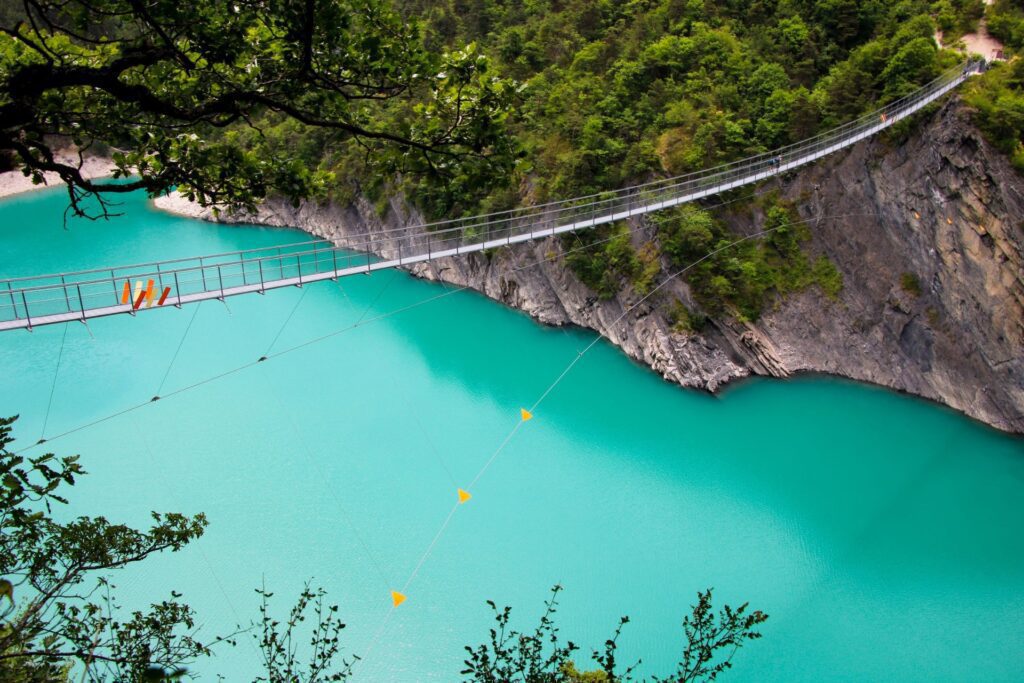 Długi, wąski most wiszący rozciągający się nad turkusowym jeziorem, otoczonym zalesionymi brzegami i skalistymi klifami. Sceneria górska.