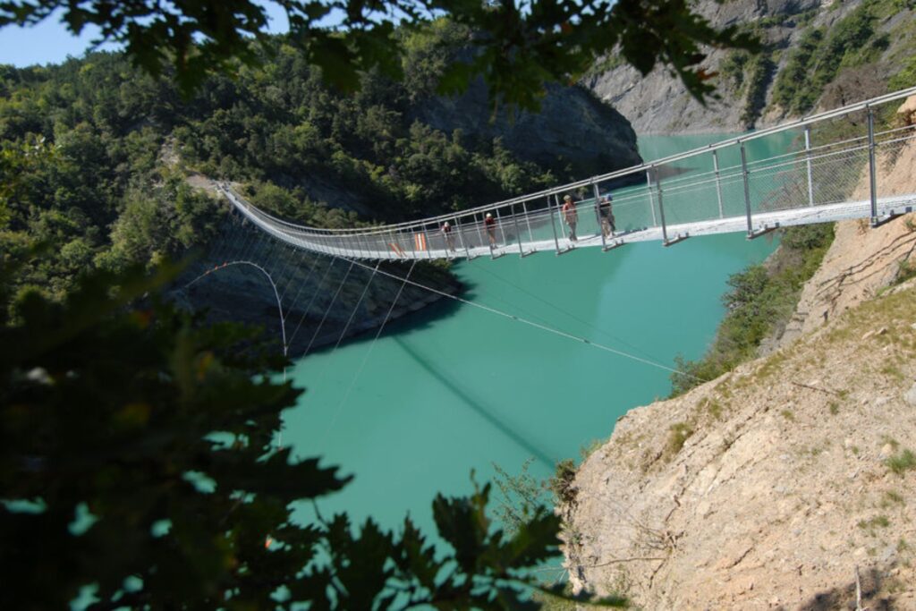 Długa kładka wisząca nad turkusowym jeziorem między zielonymi wzgórzami, z kilkoma osobami idącymi po moście w słoneczny dzień.