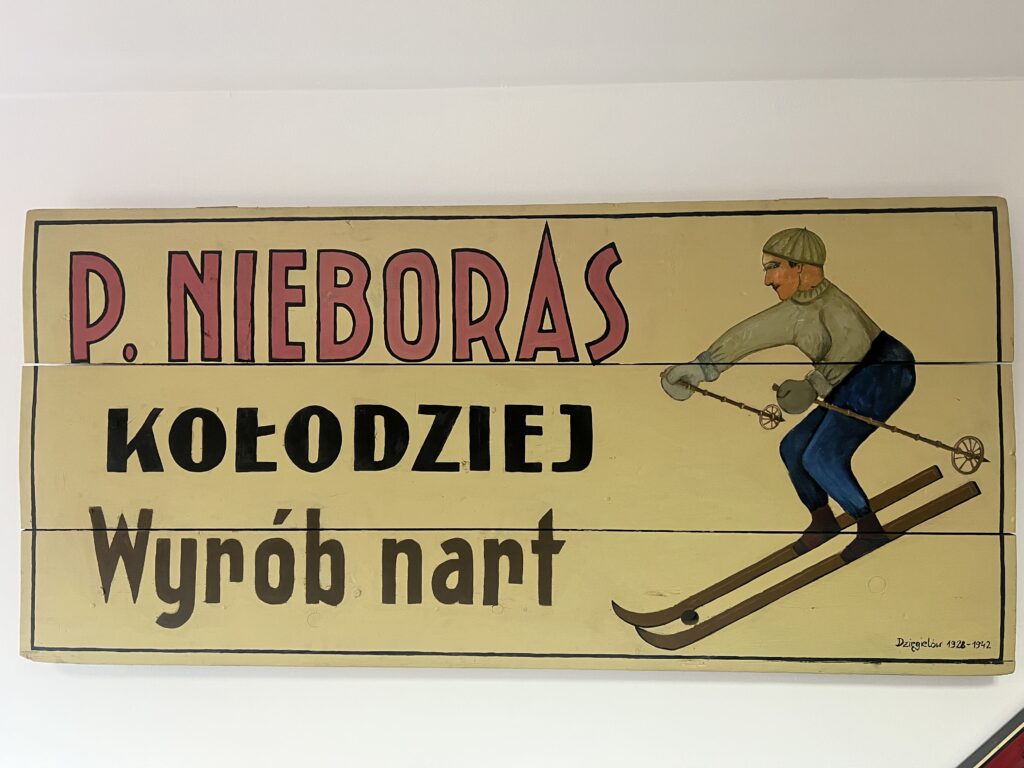 Drewniana tablica z napisem "P. Nieboras Kołodziej Wyrób nart" i rysunkiem narciarza w stylu vintage. Obraz autorstwa Dziegieciowa z lat 1938-1942.