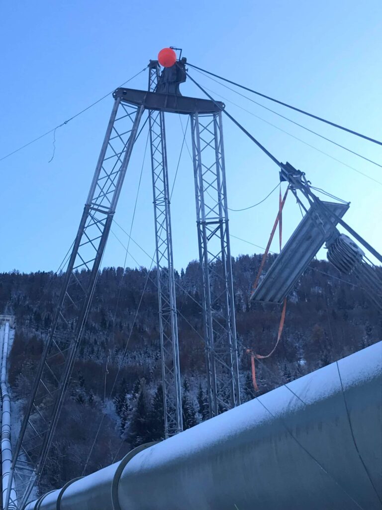 Zimowy widok na konstrukcję inżynieryjną z rurami i wysoką wieżą z pomarańczową kulą na szczycie, otoczoną zaśnieżonym lasem.