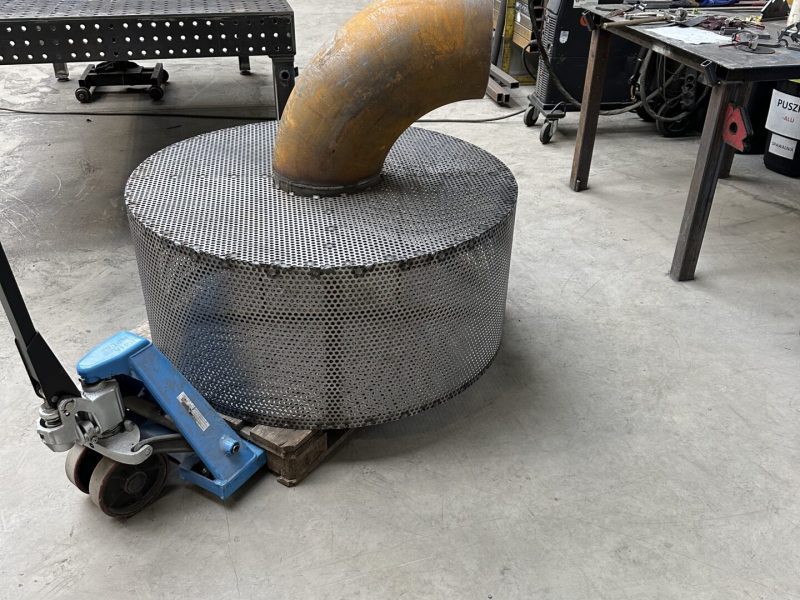 Duży metalowy filtr przemysłowy z rura na wózku paletowym w hali produkcyjnej. W tle pracownicy i sprzęt spawalniczy.