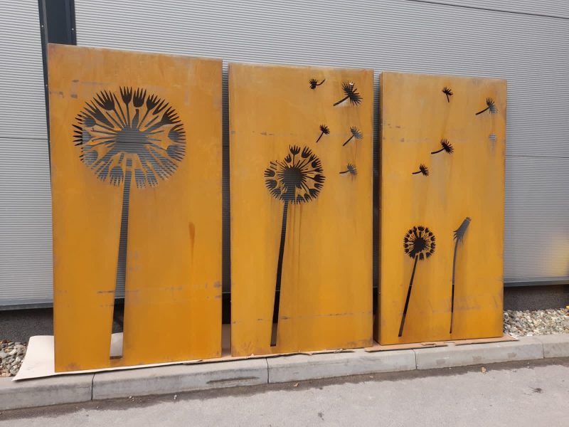 Trzy metalowe panele zardzewiałe, z wyciętymi wzorami dmuchawców w różnym stadium rozkwitu, oraz unoszącymi się nasionami. Panele stoją na zewnątrz, przed szarą ścianą.