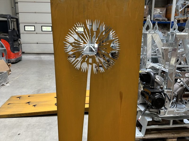 Duża, prostokątna metalowa płyta z wyciętym wzorem mniszka lekarskiego, stojąca w magazynie.